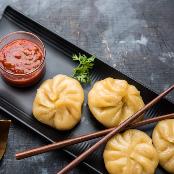 Steamed Indian momos with red chili sauce and cilantro on a black plate with chopsticks.