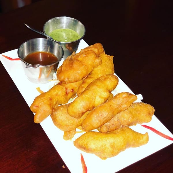 Golden-fried Indian pakora pieces on a white plate with two metal cups of dipping sauces.