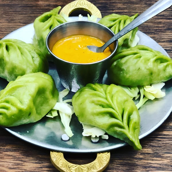 Green momos, a type of Indian dumpling, arranged around a bowl of orange dipping sauce on a metal plate.