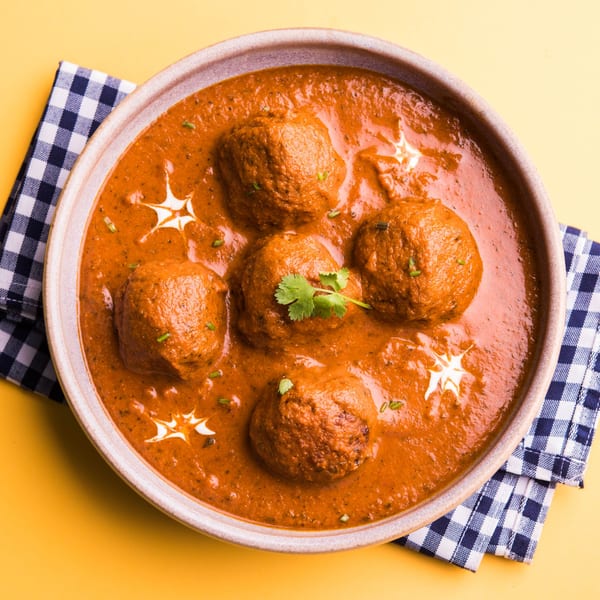 Indian Malai Kofta curry, garnished with fresh cream and cilantro, in a bowl on a yellow background.