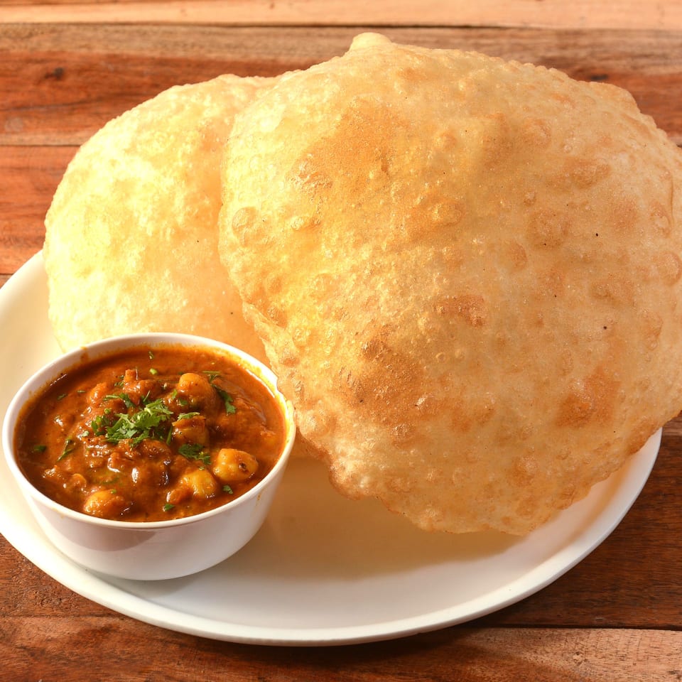 Two golden-brown Indian puri breads on a white plate with a bowl of chole masala.